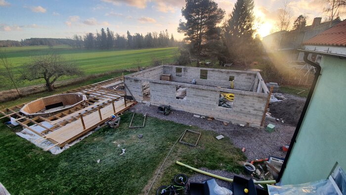 Byggarbetsplats med ett pågående garageprojekt, byggställning och takstolar, omgiven av en lantlig miljö och skog i bakgrunden.