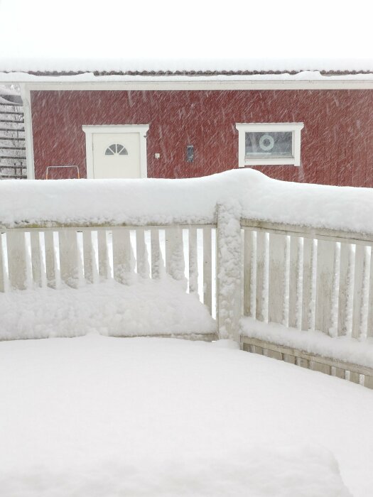 Röd byggnad i snöfall med ett vitt staket i förgrunden täckt av snö.