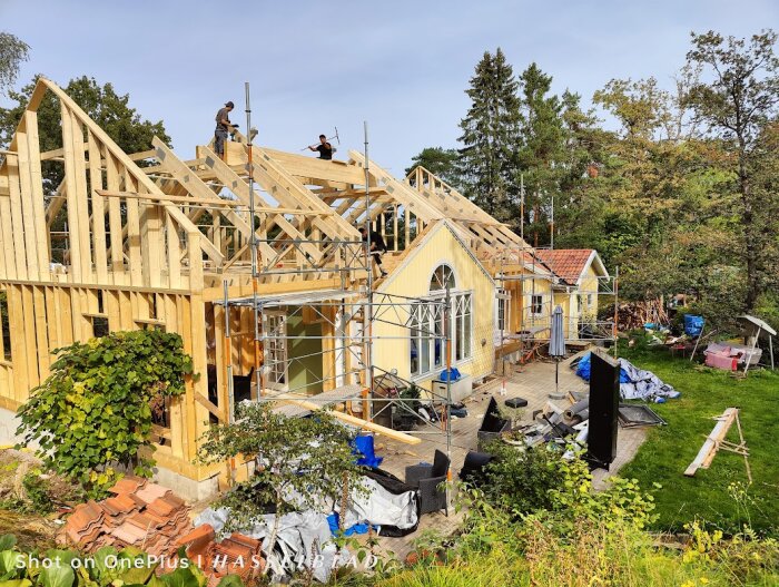 Utbyggnad av ett gult hus med nya takstolar, arbetare på taket, byggställningar och material utspridda på marken.