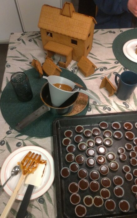 Pepparkakshus under konstruktion på ett bord, omgiven av smält choklad, redskap och en plåt med små chokladformar.