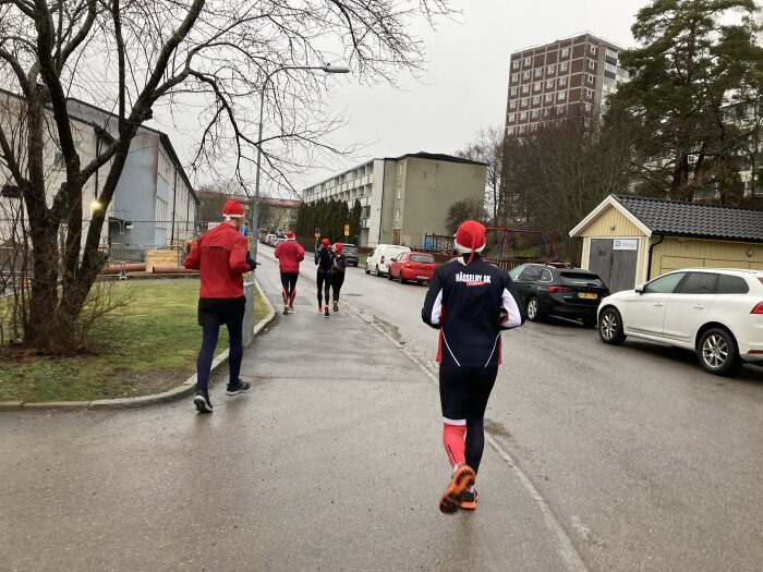 Personer joggar i tomteluvor på en regnig gata omgivna av byggnader och träd, med bilar parkerade längs vägen.