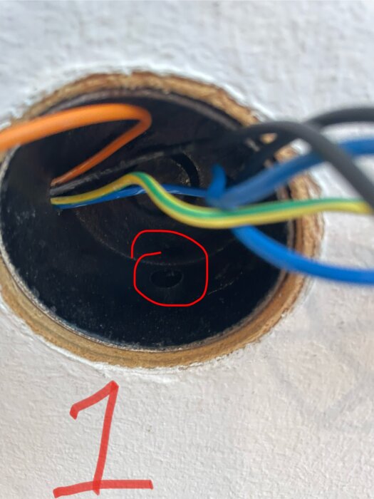 Närbild av en plåtdosa i taket med flera färgglada elkablar, varav en är orange, omgivna av spån, markerad med en röd ring och nummer 1.