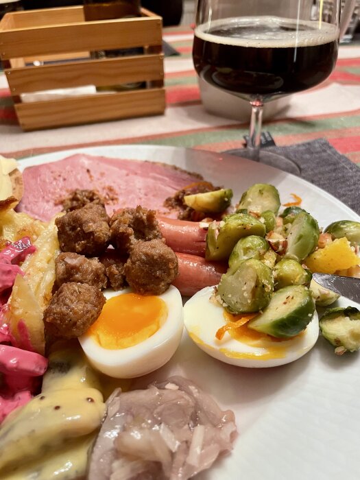 Tallrik med julmat, inklusive kokt ägg, brysselkål, köttbullar, prinskorvar och gravad lax. Ett glas mörk öl i bakgrunden.