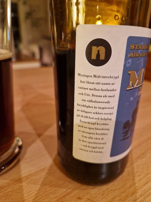 Flaska med etikett för Mysingen Midvinterbrygd, beskriver ölets inspiration och kryddighet. Står på träbord, mörkt öl i glas bredvid.
