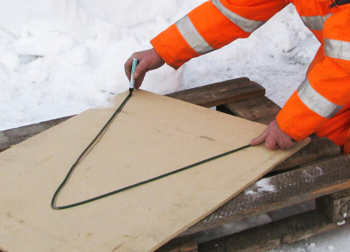 Person i orange arbetsdräkt markerar formen av ett "Peke" på en skiva med penna, utomhus i snöig miljö.