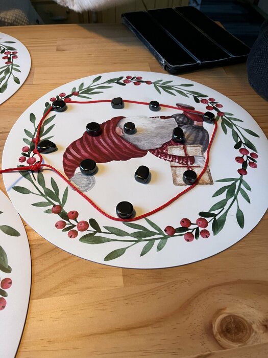 Dekoration med svart stenar och rött snöre på rund tallrik med julmotiv av tomte och blommor.