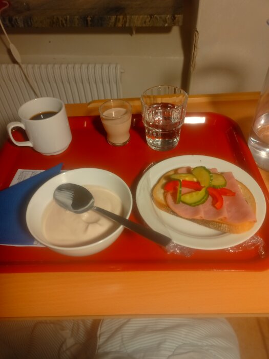 Frukostbricka med yoghurt, smörgås med skinka och grönsaker, kaffe, juice, och vatten på ett rött plastfat.