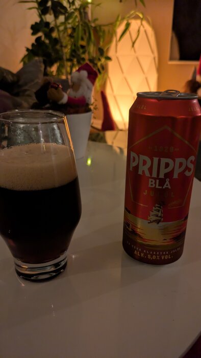 Ett glas med skummande mörk dryck bredvid en Pripps Blå julölburk på ett bord med växter och juldekorationer i bakgrunden.