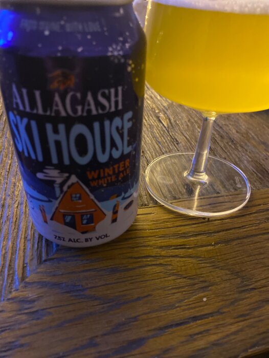 Burk med Allagash Ski House Winter White Ale och ett halvtomt glas på ett träbord.