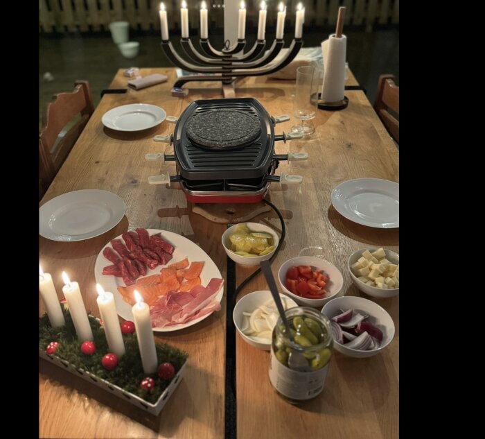 Dukat bord med raclettegrill, tallrikar med kött och ost, samt skålar med grönsaker. Ljusstake i bakgrunden.