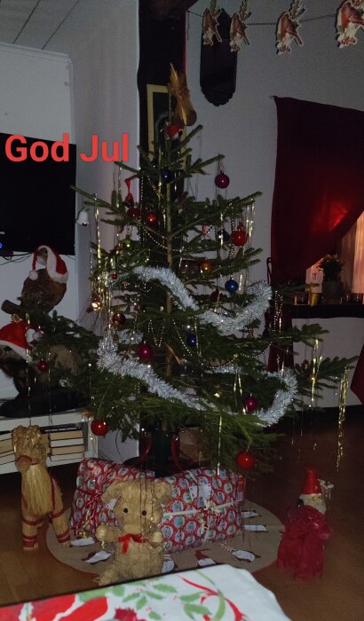 Julgran dekorerad med kulor och glitter, omgiven av julklappar och halmfigurer. Text "God Jul" syns på bilden.