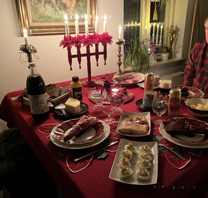 Julbord med levande ljus, jultallrikar, ägghalvor, lax, ostar och drycker på rött dukat bord i mysig hemmamiljö.