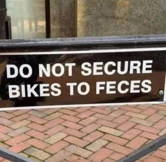 Skylt med texten "DO NOT SECURE BIKES TO FECES" framför en trottoar av marksten.