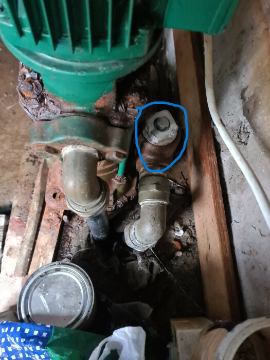 Närbild av en gammal Grundfos ejektorpump som visar en fastrostad grå plugg markerad med blå cirkel, omgiven av rör och rostiga ytor.