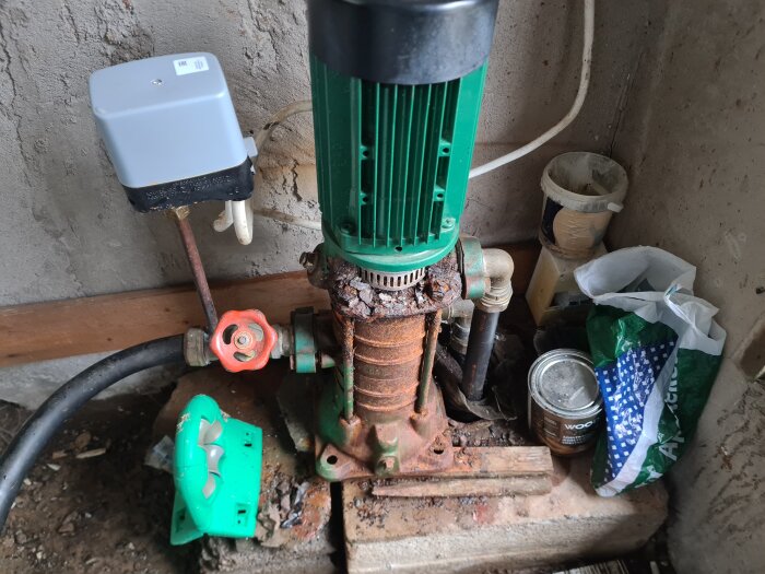 Rostig Grundfos CPE 3-60 ejektorpump med röd ventil och borrade hål, omgiven av verktyg och påsar, installerad på cementblock i ett förråd.