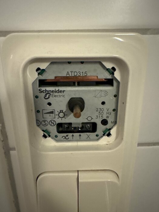 Närbild av en Schneider Electric dimmer modell ATD315, monterad på en vit kakelvägg, med synliga justeringsknappar och märkningar.