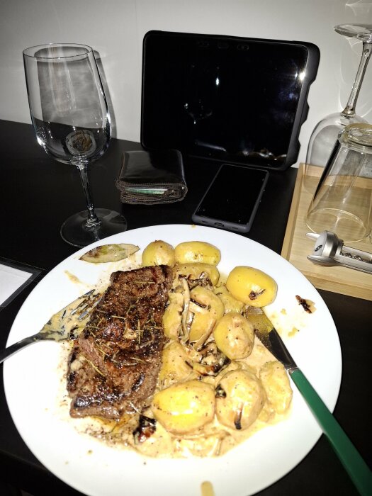 En tallrik med entrecote och gräddstuvad potatis på ett skrivbord, bredvid ett vinglas, en plånbok och en surfplatta.