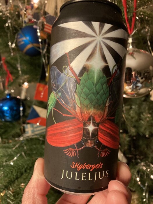 En hand håller en burk med etiketten "Stigbergets Juleljus", framför en julgran med dekorationer i bakgrunden.