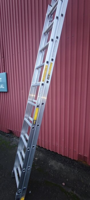 Aluminiumstege lutad mot en röd korrugerad vägg, märkt som prisvärd.