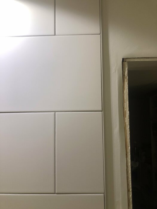 Oregelbundna vita kakelplattor med ojämn fogbredd på en vägg, med synlig dörröppning till höger.
