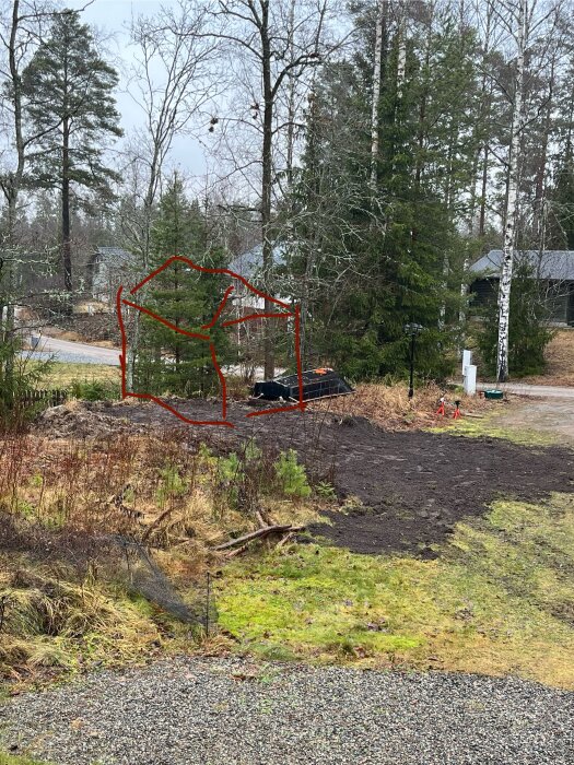 Skogslandskap med upphöjning och röd markerad framtida plats för friggebod nära träd och växter. Synlig grusväg i bakgrunden.