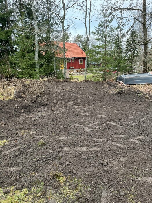 Upphöjd jordyta med skog i bakgrunden. En friggebod ska placeras här på en grund av makadam. Rött hus med tegeltak i bakgrunden.