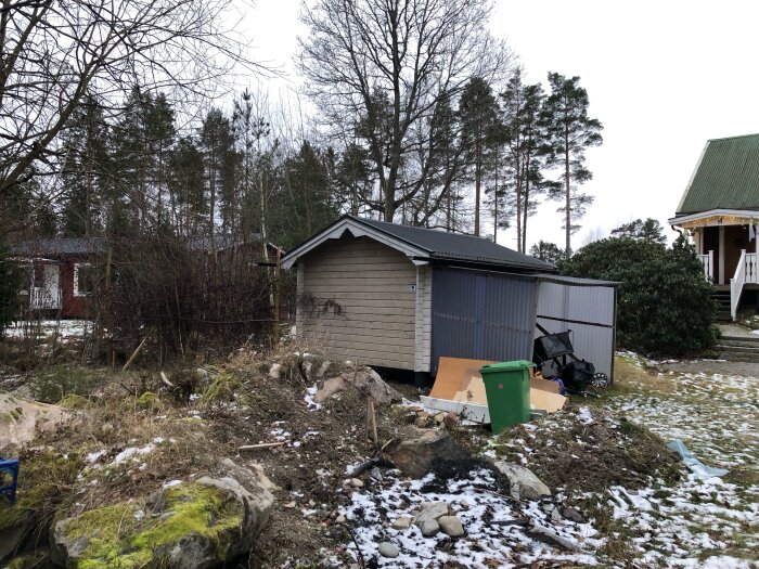 En friggebod står vid en snötäckt tomt med olika föremål utspridda runtom, omgiven av träd och en annan byggnad synlig i bakgrunden.