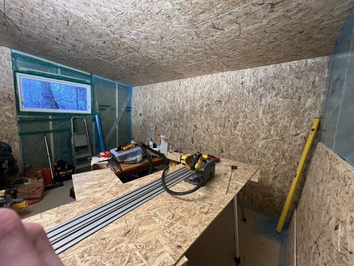 Byggrum med OSB-skivor på väggar och tak, arbetsbord med verktyg och en såg, stege vid fönster, plastöverdrag och byggmaterial i bakgrunden.
