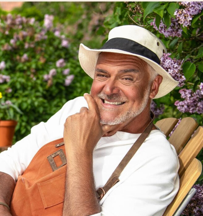 Man i hatt och förkläde ler medan han sitter utomhus omgivet av blommor och grönska.