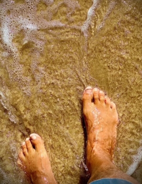 Fötter på sandstrand med vågor sköljande över tårna.