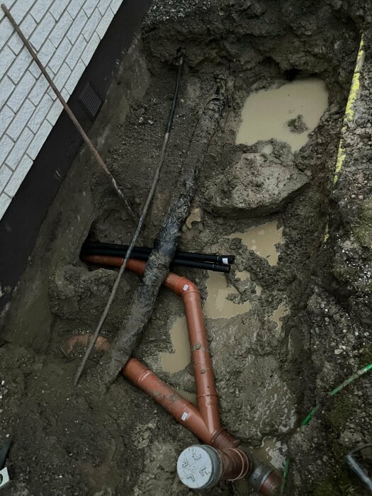 Ny grävd grav med anslutna avloppsrör utanför ett hus, del av arbete för nytt badrum. Marken visar lera och vattenpölar.