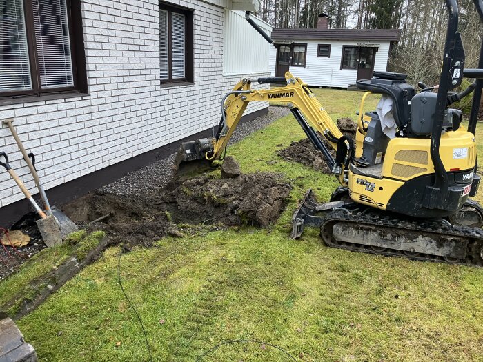 Minigrävare i arbete utanför ett hus, grävning för ny stambytesinstallation. Tydlig jordhög bredvid och spadar vid sidan av väggen.