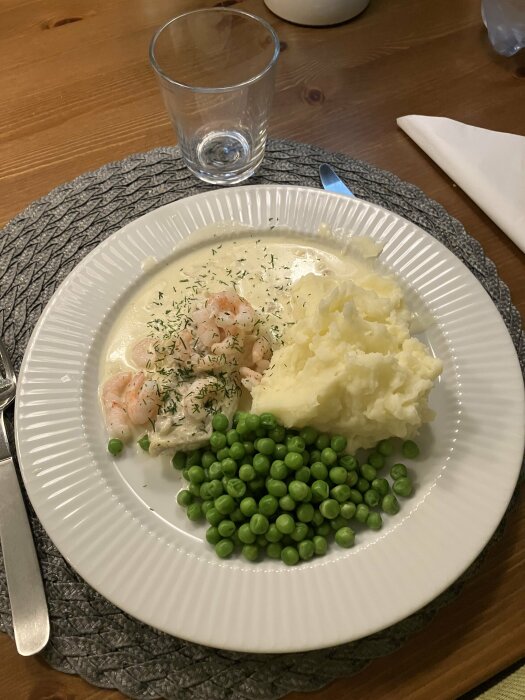 Torskrygg med räkor i sås, serverad med potatismos och gröna ärtor på en tallrik, dukad med glas och bestick på träbord.