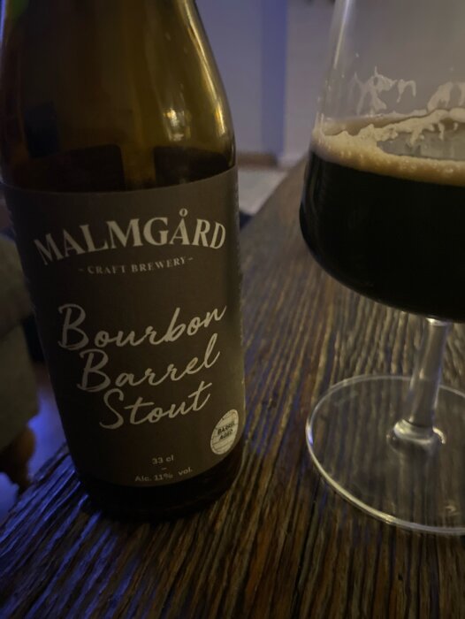 Flaska Malmgård Bourbon Barrel Stout och ett glas med mörk stout på ett trägolv.