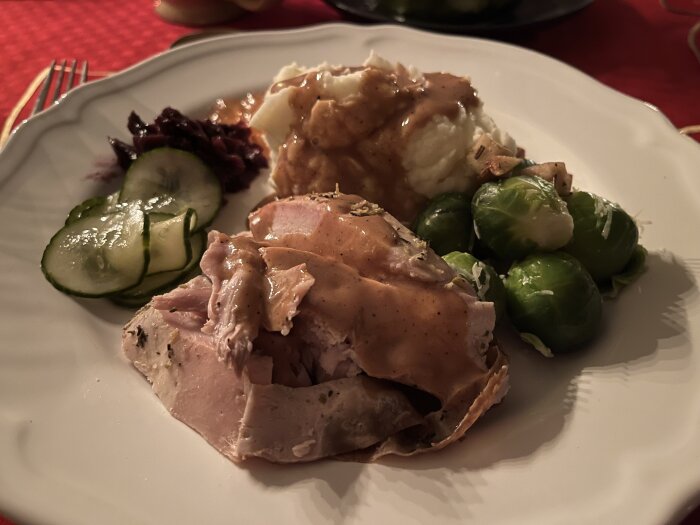 En tallrik med kalkon, potatismos med sås, brysselkål, inlagd gurka, rödkål och äppelfyllning på ett dukat bord.