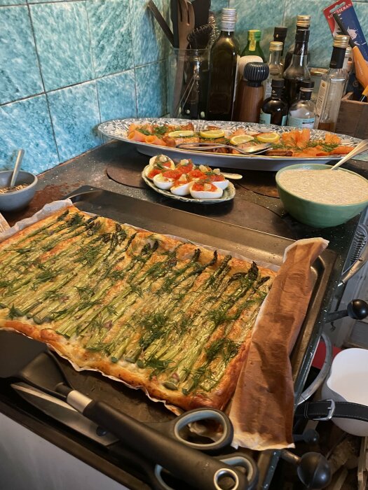 Grönt kakel i kök med bricka med mat, inklusive sparrispaj och ägghalvor, samt flaskor och köksredskap i bakgrunden.