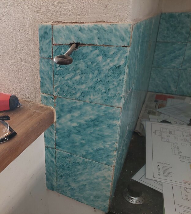 Närbild på blågröna kakelplattor med ojämnt mönster, monterade i ett hörn. En metallkrok är fastsatt inuti en kakelfog.