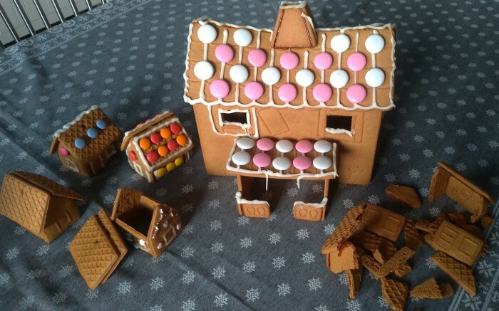 Pepparkakshus under konstruktion med färgglada chokladlinser som detaljer på taket, omgivna av små pepparkaksdelar och ett snötäcke som duk.