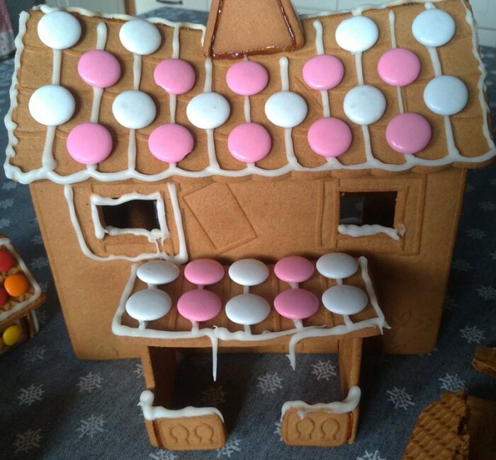 Pepparkakshus dekorerat med vita och rosa knappar och glasyr på tak och veranda, stående på en grå duk med stjärnmönster.