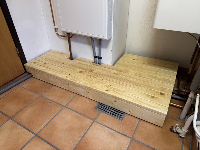 Träplattform i garage, skyddar rör och fungerar som steg; gjord av ljusa träplankor ovanpå kakelgolv.
