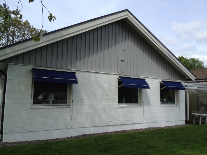 A white brick house facade with three windows, each having a blue awning. The house is surrounded by grass and has a stone border at the foundation.