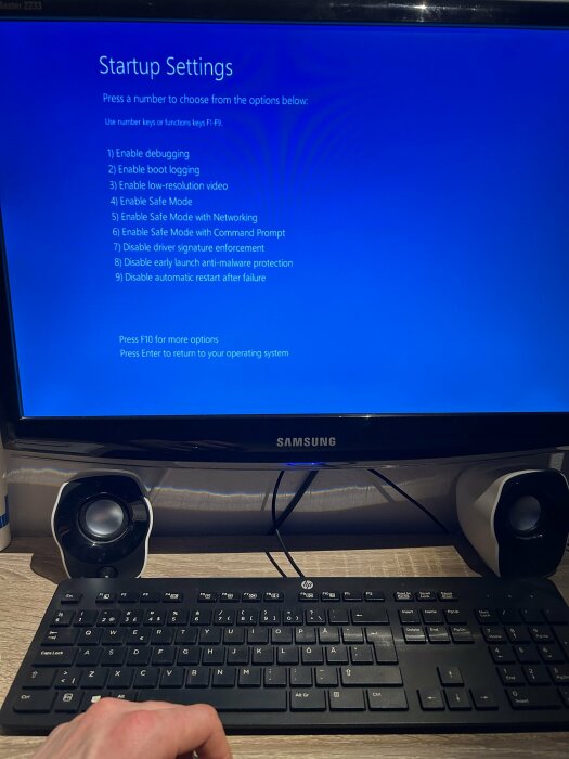 Datorskärm visar "Startup Settings" med alternativ för felsäkert läge och andra startinställningar. Tangentbord och högtalare syns.