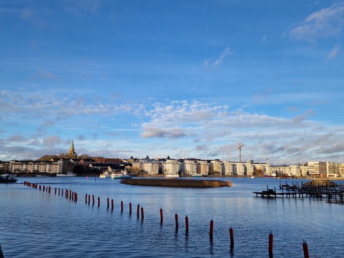 Stadsbild av bebyggelse vid vattnet med en kyrktorn synligt, blå himmel och moln i bakgrunden, pålar syns i vattnet.