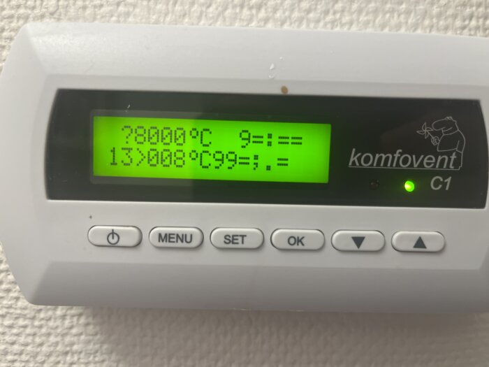 Digital termostatdisplay som visar oklara tecken där temperaturen väntas. Text "komfovent C1" synlig, med knappar på nederdelen av enheten.