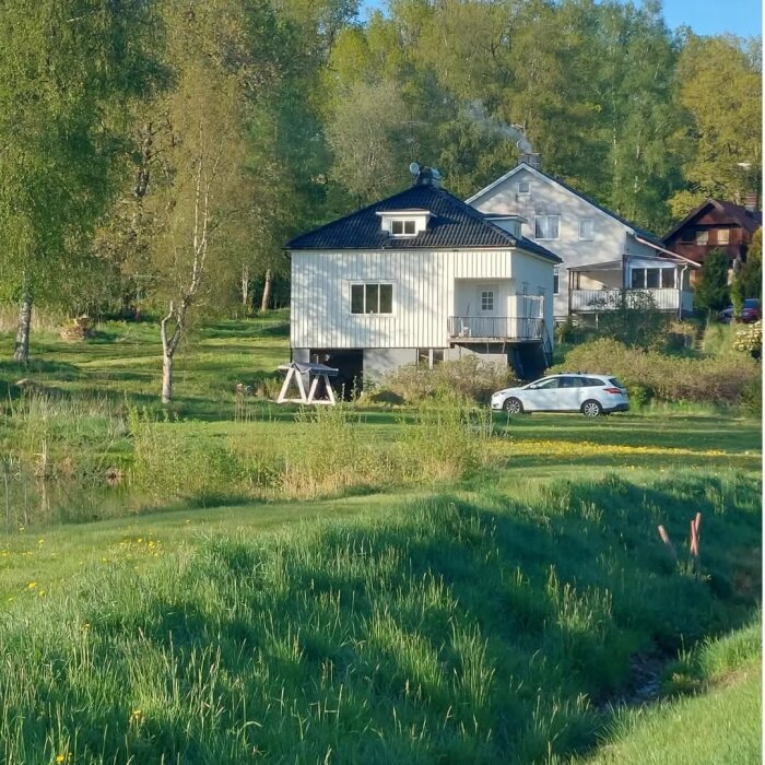 Vitt hus med pyramidtak och fönsterkåpor i en grönskande miljö. Parkerad bil syns framför huset. Skog och gräsmatta omger byggnaden.