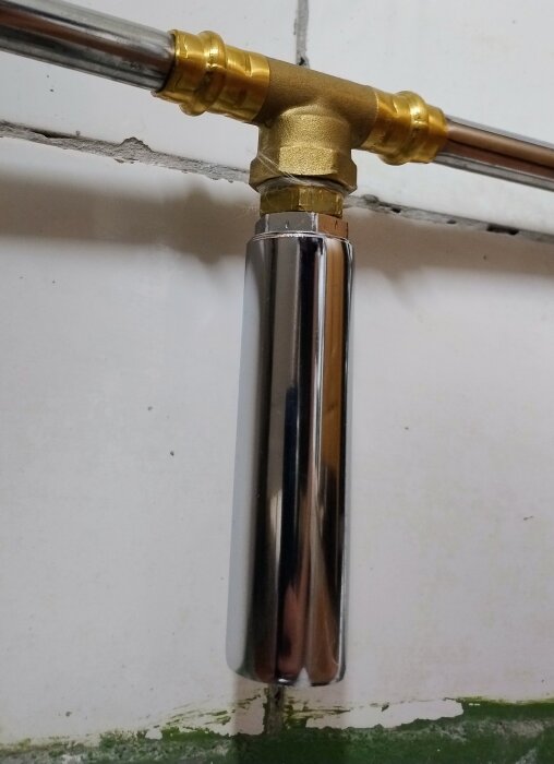 Kromfärgad dämpare monterad på en röranslutning i mässing mot en kaklad vägg.