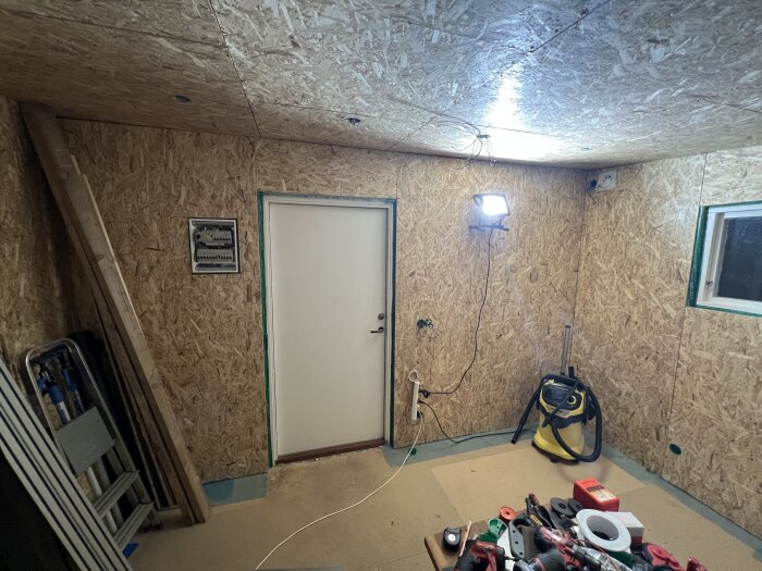 Rum med OSB-klädda väggar, arbetslampa, bord med verktyg och en dammsugare. Färdigt första lager, redo för gipsning.