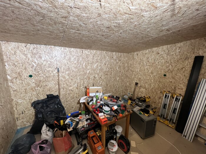 Stökigt rum under renovering med träklädda innerväggar och ett bord fullt av verktyg och material. Stegar och skräp syns också på golvet.