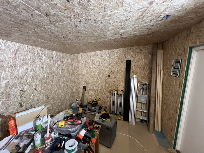 Rörigt arbetsbord med verktyg och material i ett rum med första lagret på innerväggarna klart; stegar och brädor lutar mot väggen.