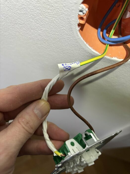 Hand som håller elektrisk kabel med märkning, visas framför en vägguttag med flera ledningar, inklusive en jordad gröngul kabel.